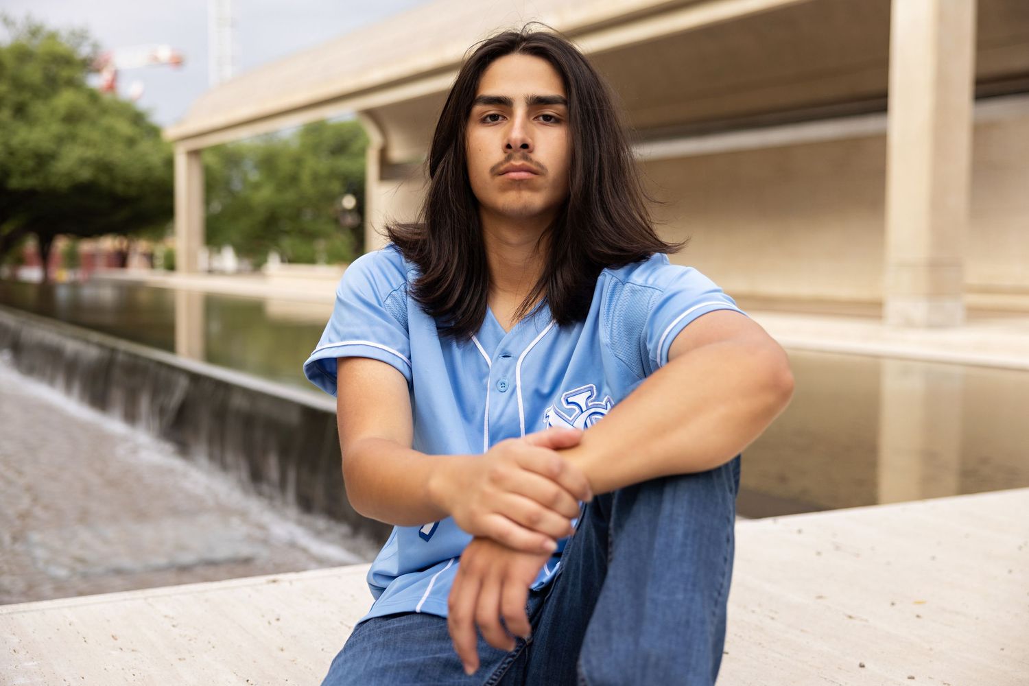 Young man with long dark hair wearing a blue shirt sits outdoors near a water feature in an urban setting.