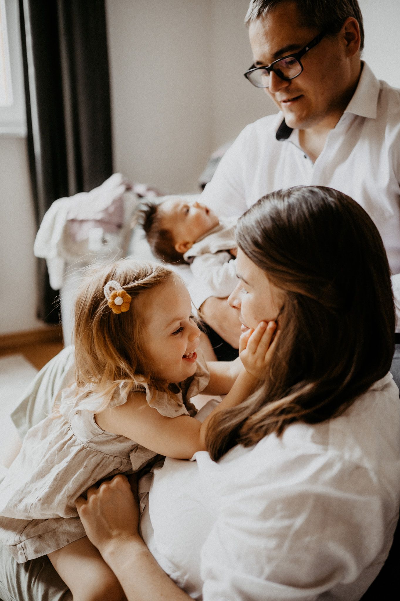 A warm family moment with parents and two children, including a newborn, sharing tender embraces in a softly lit room.