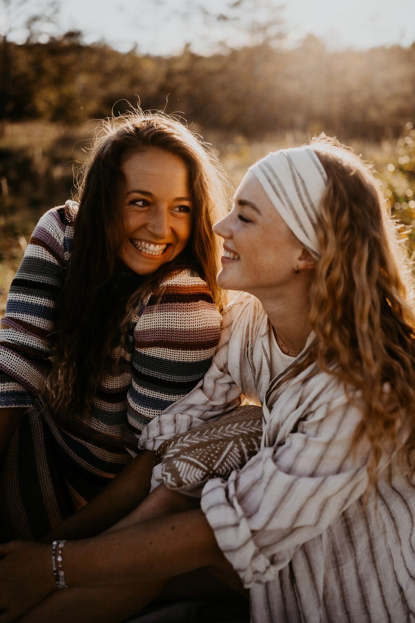 Two smiling young women sitting outdoors in golden sunlight, wearing casual striped outfits, sharing a joyful moment together.