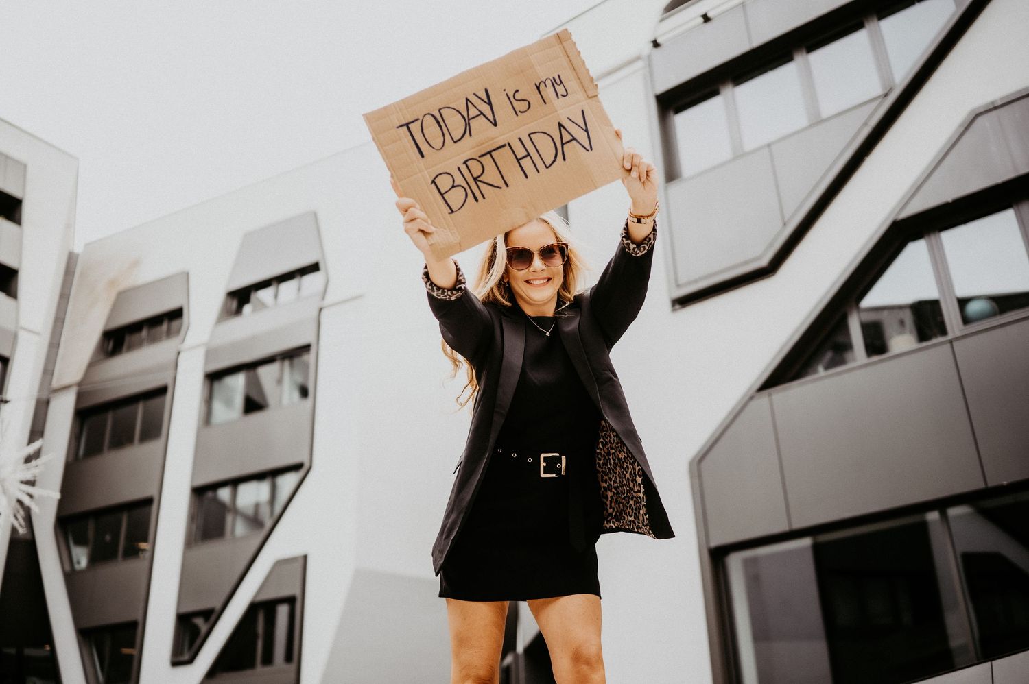 Woman holding a cardboard sign reading Today is my Birthday, smiling outdoors near a modern building.