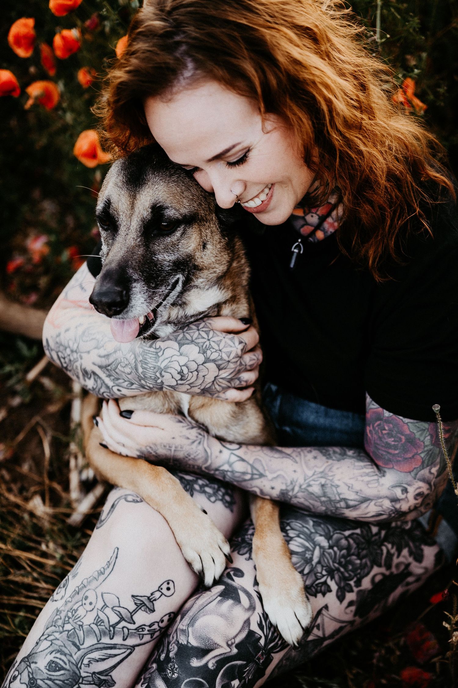 Smiling redhead woman with tattoos hugging a German Shepherd dog among orange flowers outdoors.