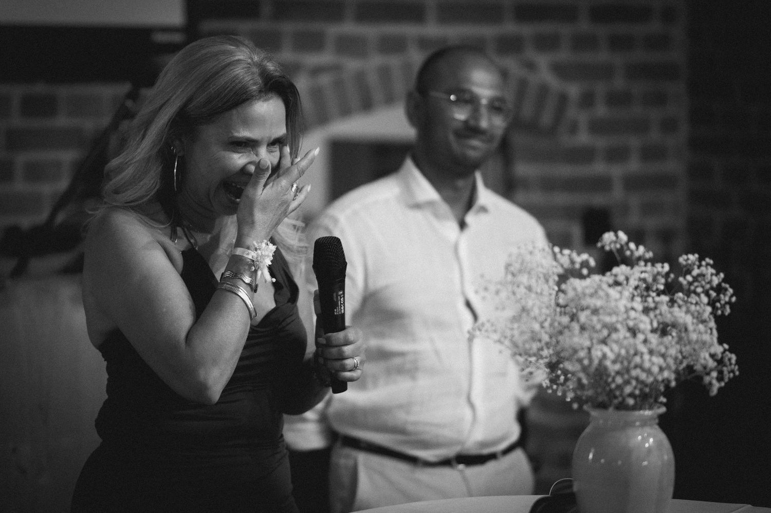 Black and white photo of a woman laughing at a microphone beside a smiling man near a floral arrangement.