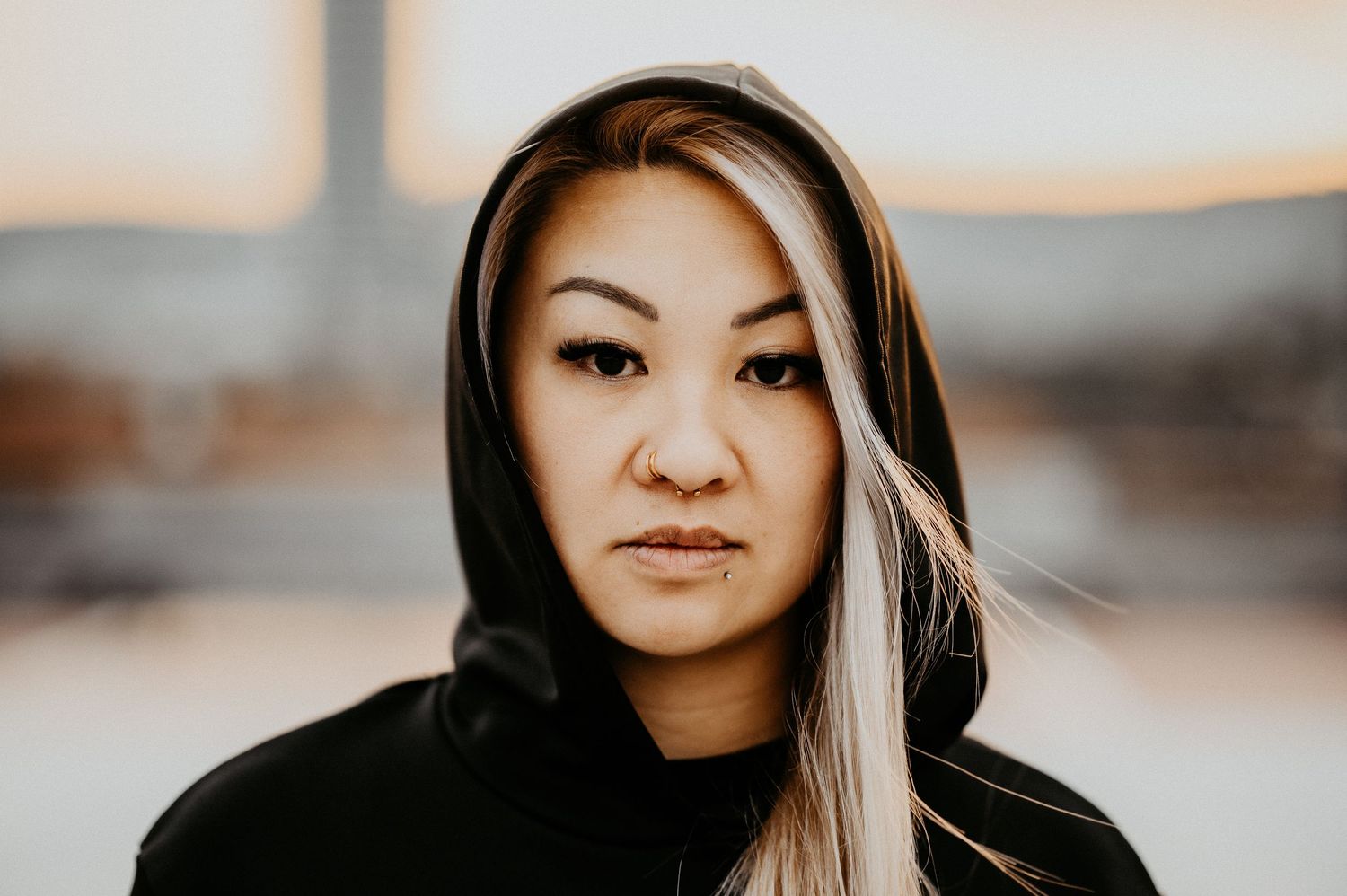 Young Asian woman in black hoodie with blonde highlights poses outdoors at sunset with serious expression.