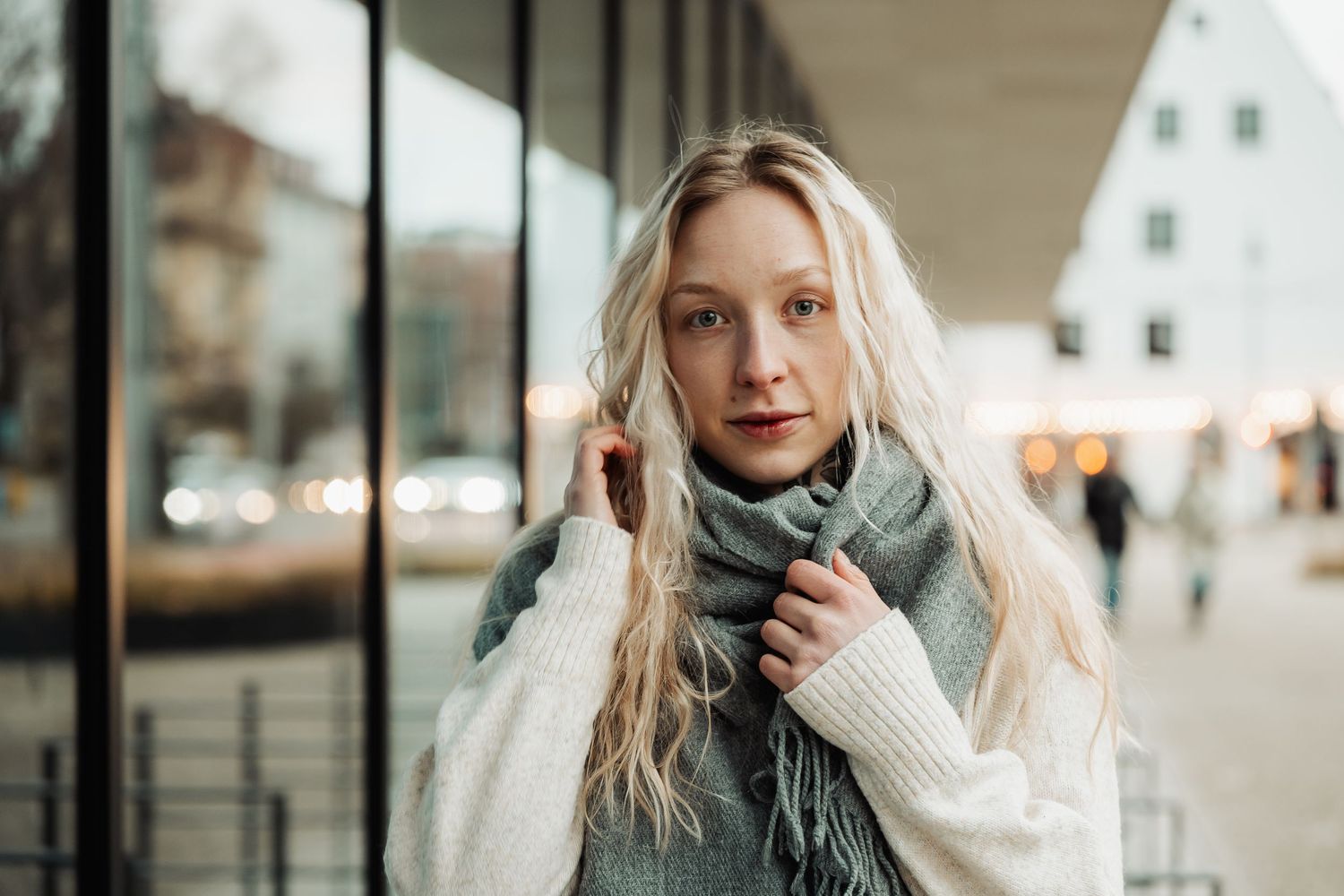 Young blonde woman in white sweater and gray scarf standing outdoors in urban setting during winter.