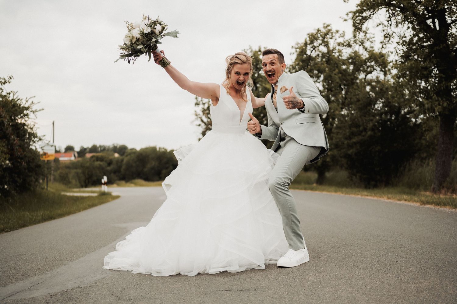 Joyful newlyweds celebrating on a road, bride in white gown holding bouquet, groom in gray suit, both laughing.