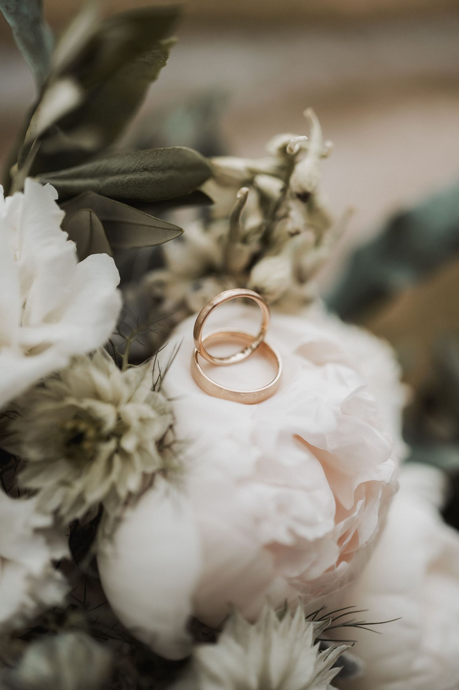Two gold wedding bands resting on white peony blooms in an elegant bridal bouquet with greenery.