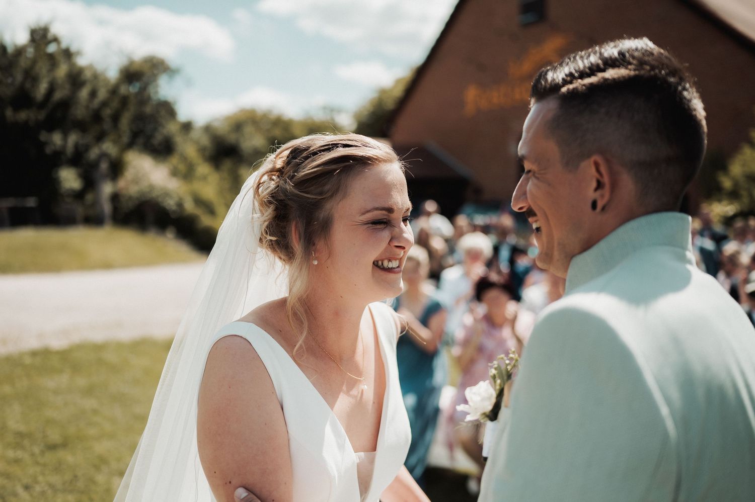 Joyful bride in white gown and veil laughing with groom in mint suit at outdoor wedding ceremony.
