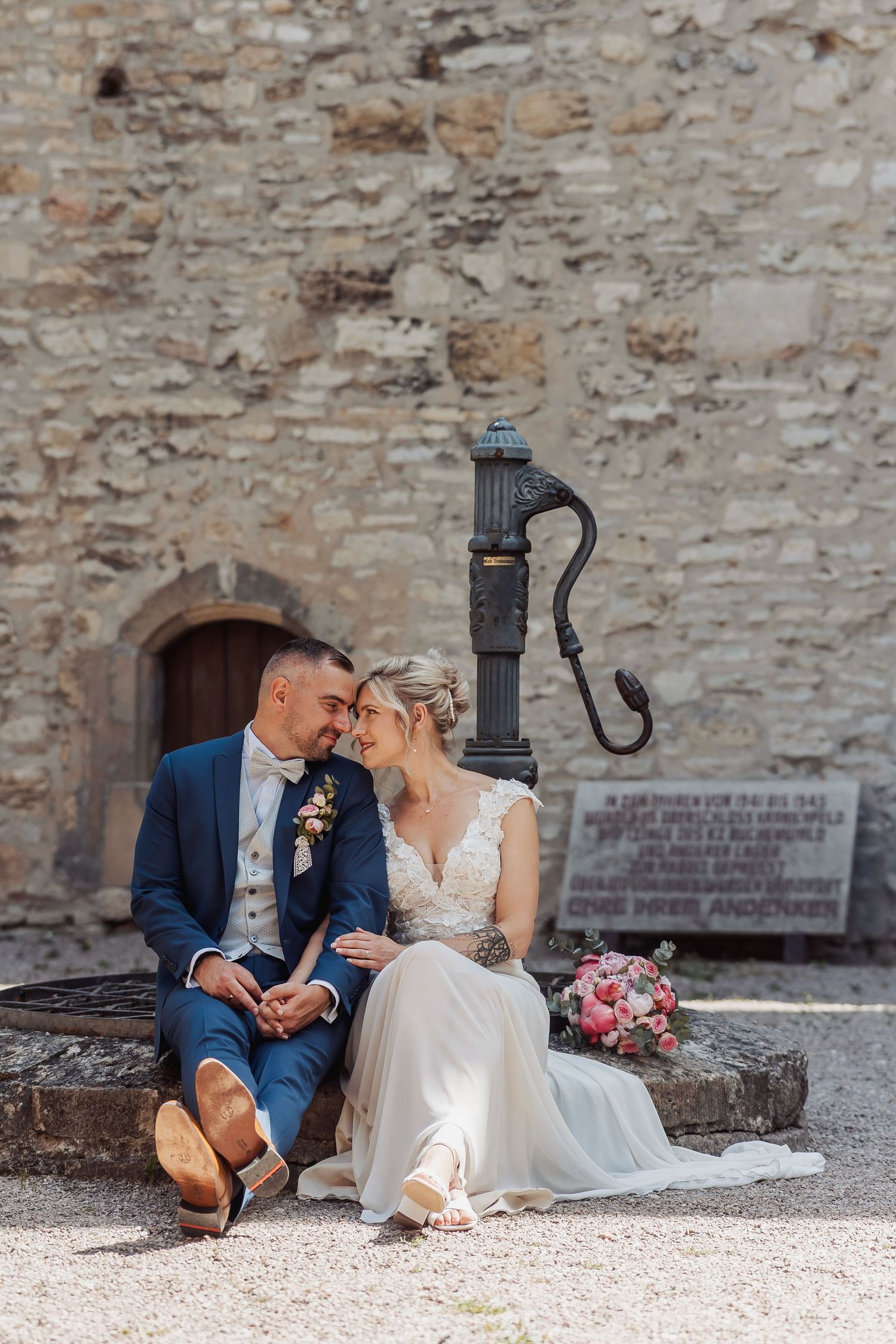 Bride and groom sit together by a vintage stone water pump, sharing an intimate moment in their wedding attire.