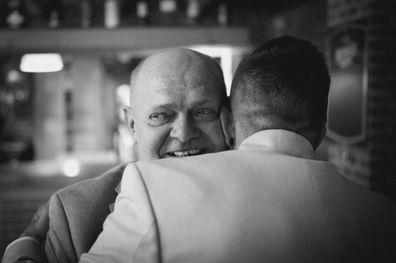 Two men sharing a warm embrace, smiling joyfully in a black and white candid photo at an indoor event.
