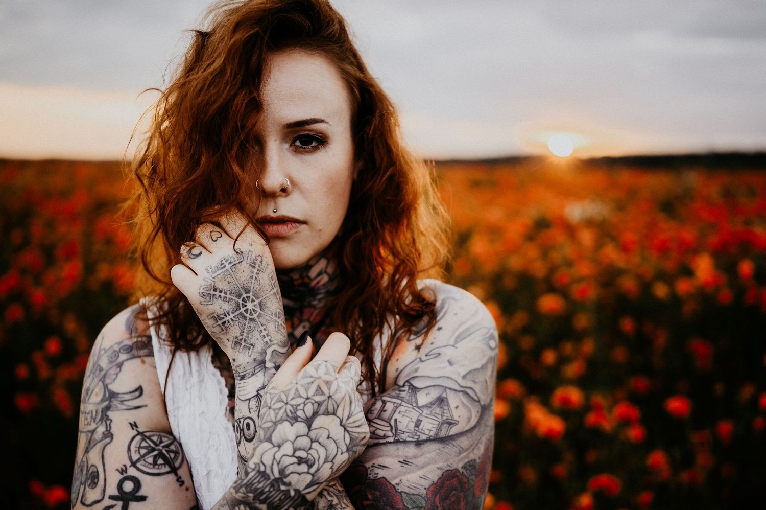 Tattooed woman with auburn hair poses in a vibrant poppy field at golden sunset, arms crossed over chest.