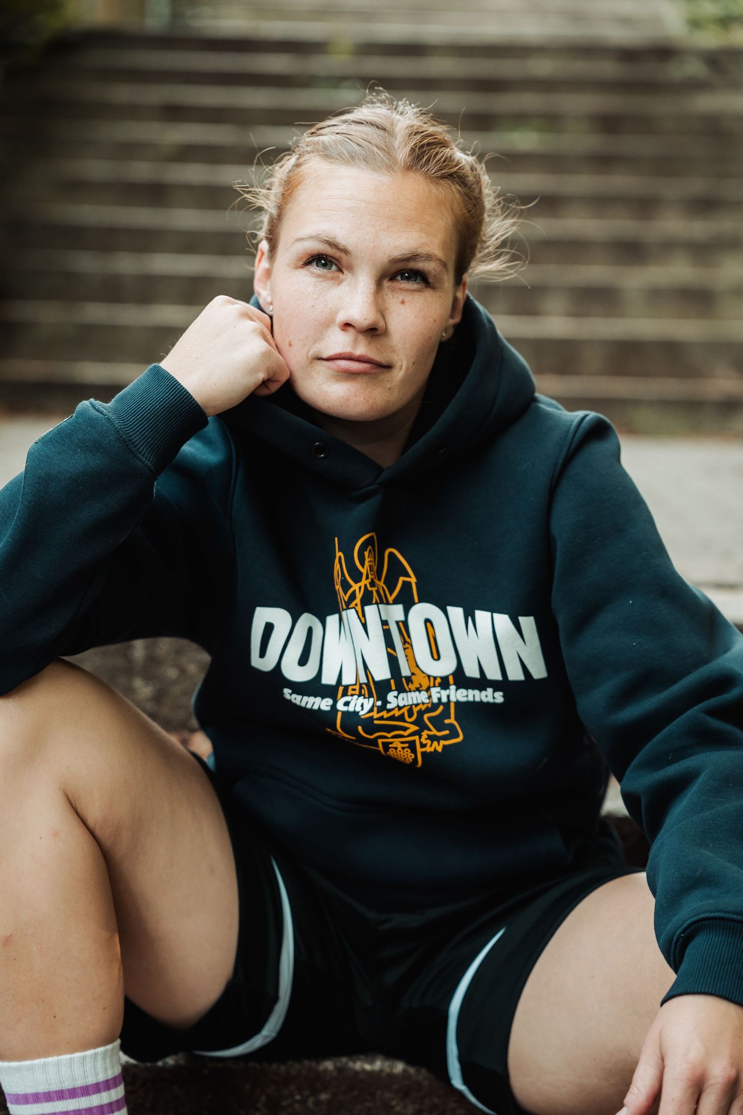 Woman sitting outdoors wearing a dark green Downtown hoodie and shorts, with stairs in the background.