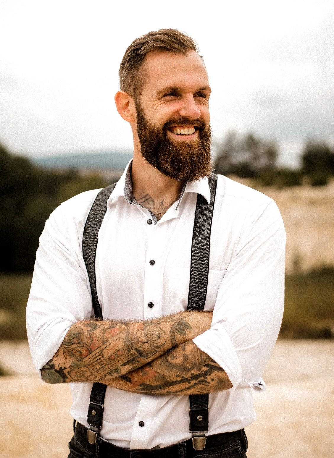 Smiling bearded man with tattoos wearing white shirt and suspenders standing outdoors with arms crossed.