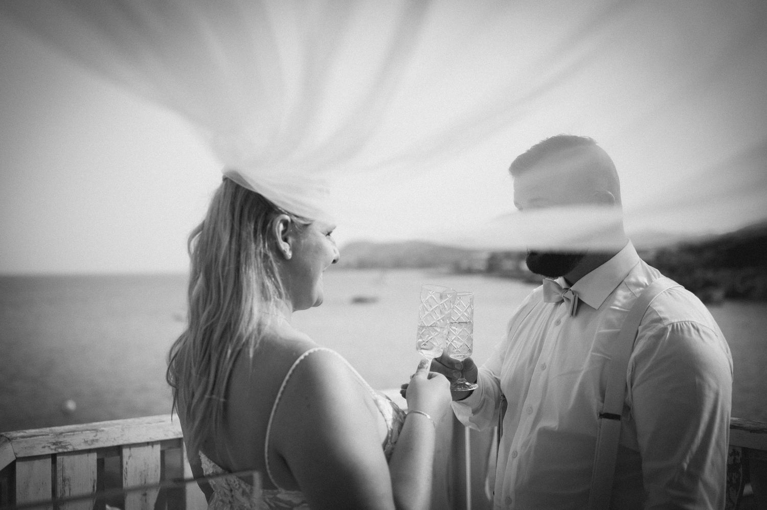 Black and white photo of a couple toasting with champagne glasses on a balcony overlooking the ocean.