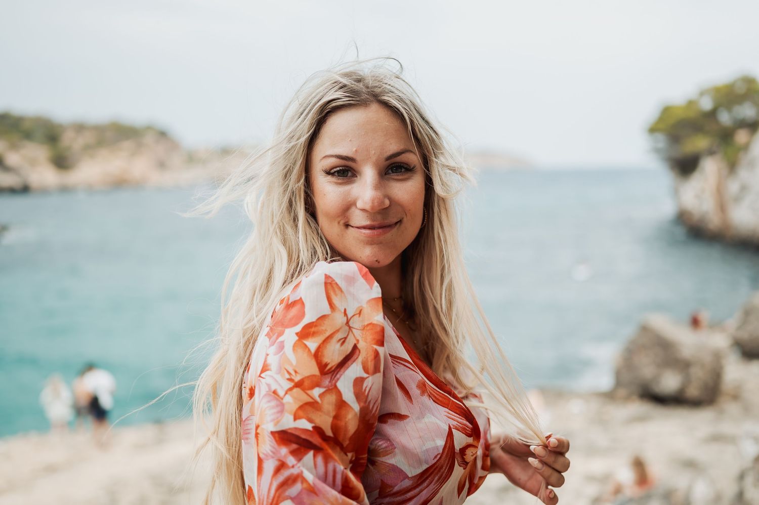 Smiling blonde woman in floral dress poses on rocky coastal cliffs with turquoise sea in background.
