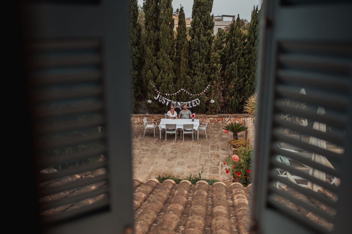 View through shuttered window of an outdoor wedding reception table with Just Married banner among tall cypress trees.