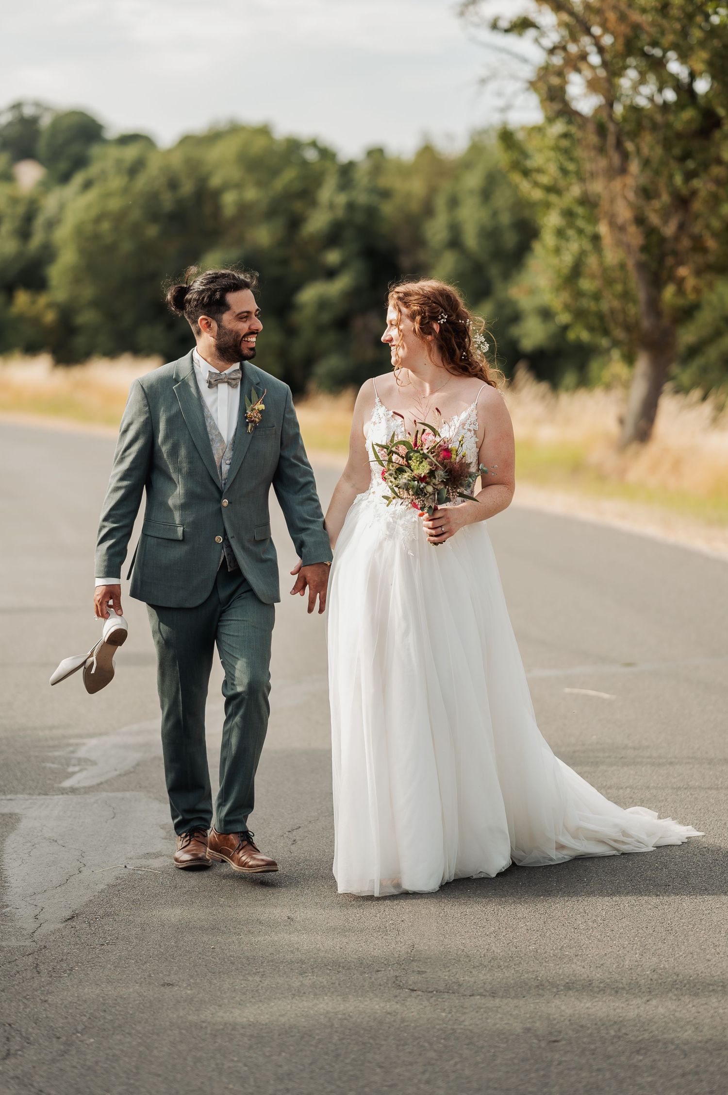 A bride and groom walk hand in hand on a road, she holds a bouquet, he carries her shoes, surrounded by trees.