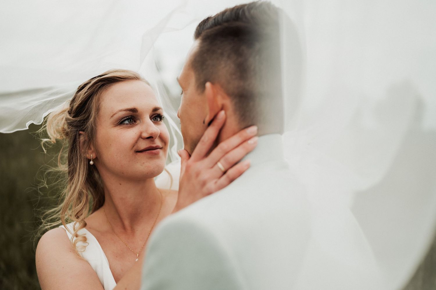 Bride gazing lovingly at groom under flowing white veil, wearing wedding ring, romantic outdoor wedding portrait.