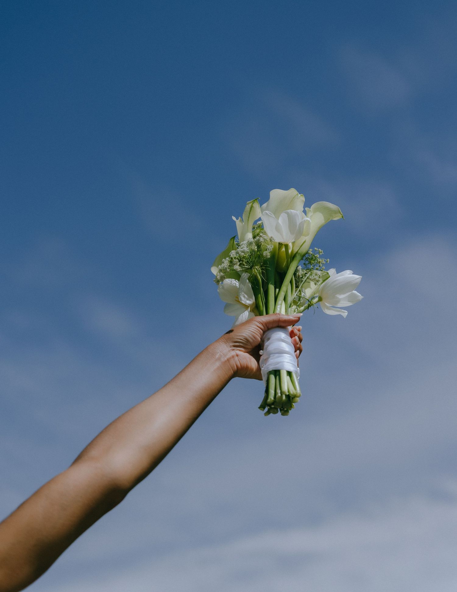 Hand holding up a white flower against a blue cloudy sky background.