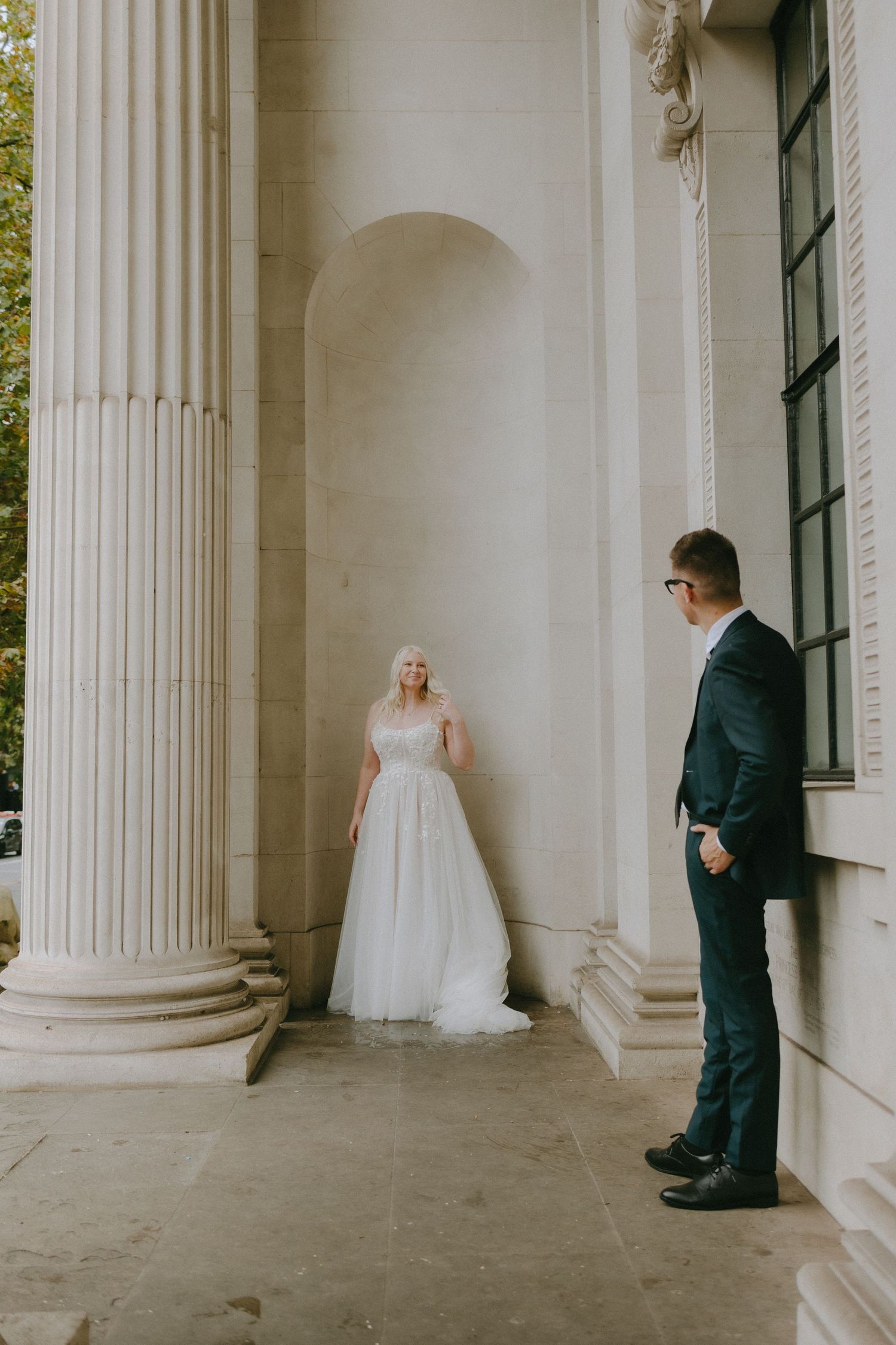 A bride in a white ball gown and groom in dark suit stand between elegant white columns at a historic venue.