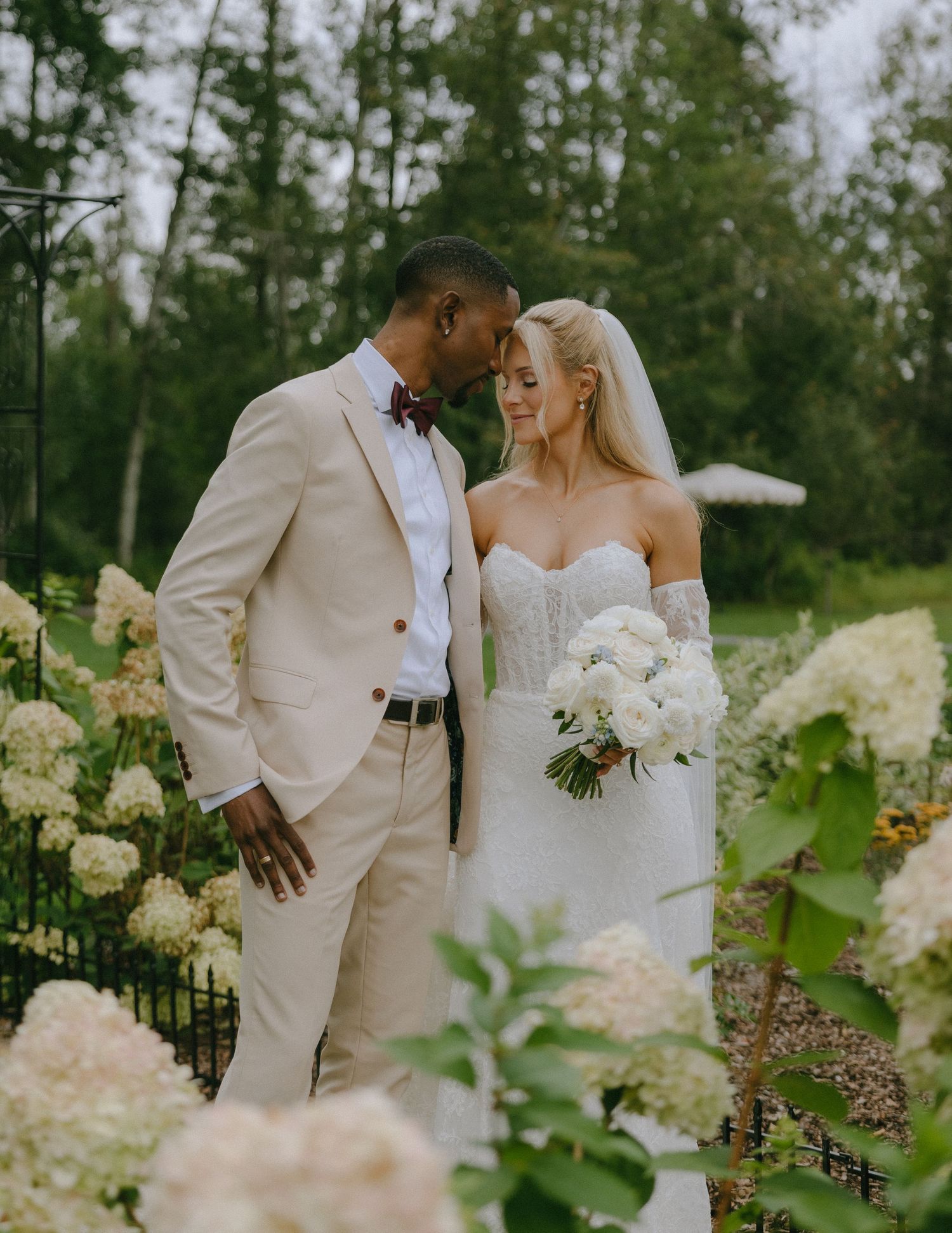 A wedding couple shares an intimate moment outdoors surrounded by white hydrangea blooms and greenery.