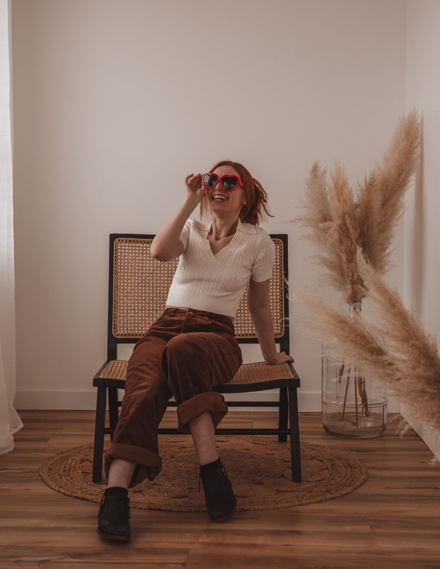 Portrait of someone in sunglasses and white top posing on a rattan chair with pampas grass decor.
