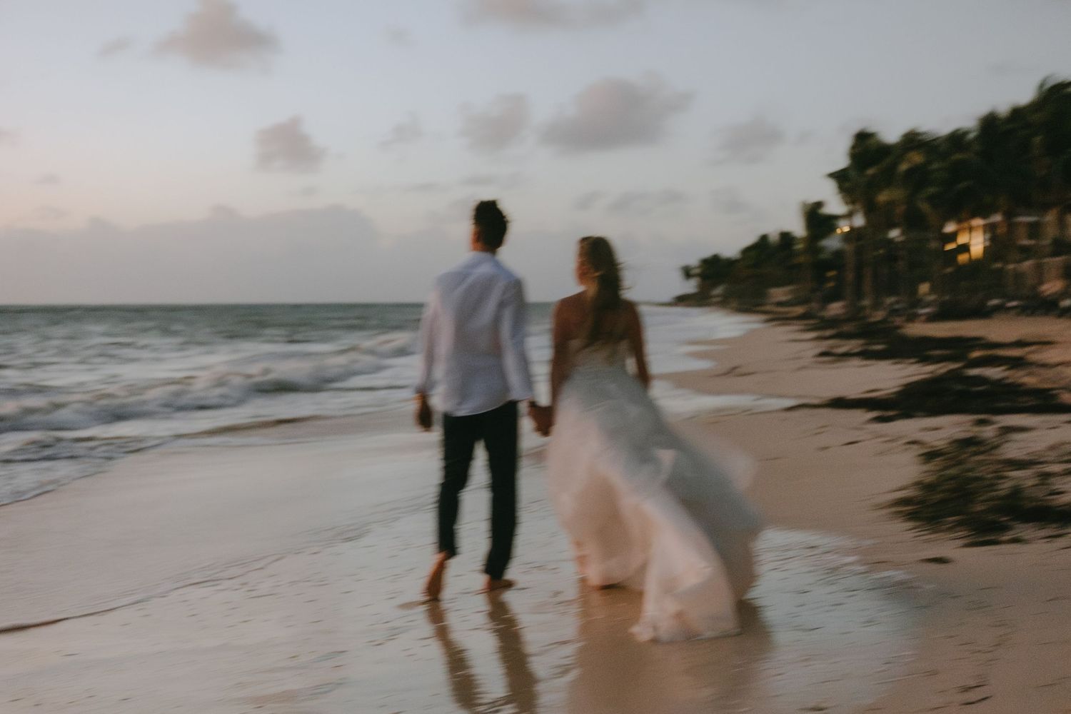 Two people walk hand in hand along a scenic tropical beach at sunset with palm trees and houses in the distance.