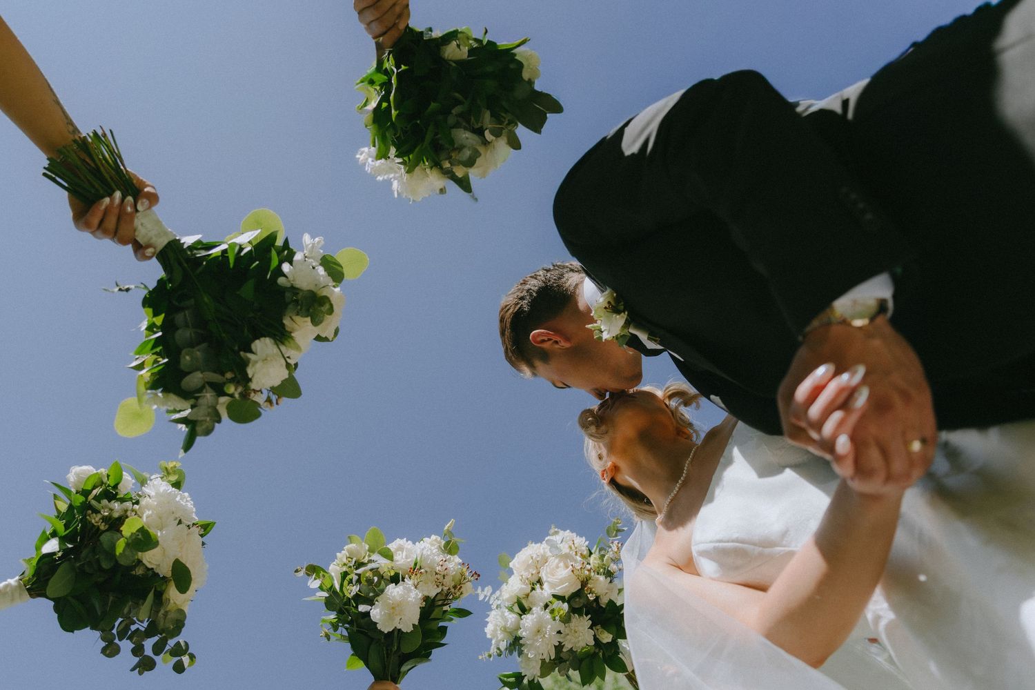 Low-angle view of bride and groom kissing surrounded by wedding party holding white floral bouquets against blue sky.