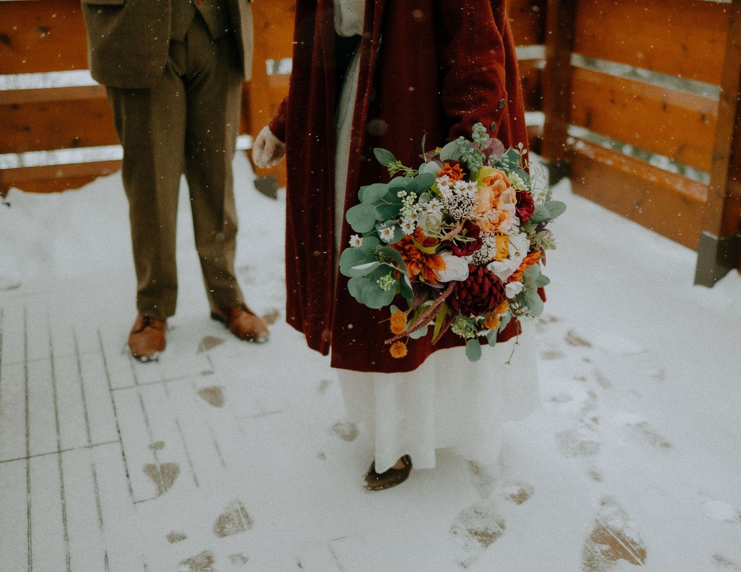 A winter bouquet of peach and burgundy flowers with eucalyptus held against a snowy background and red coat.