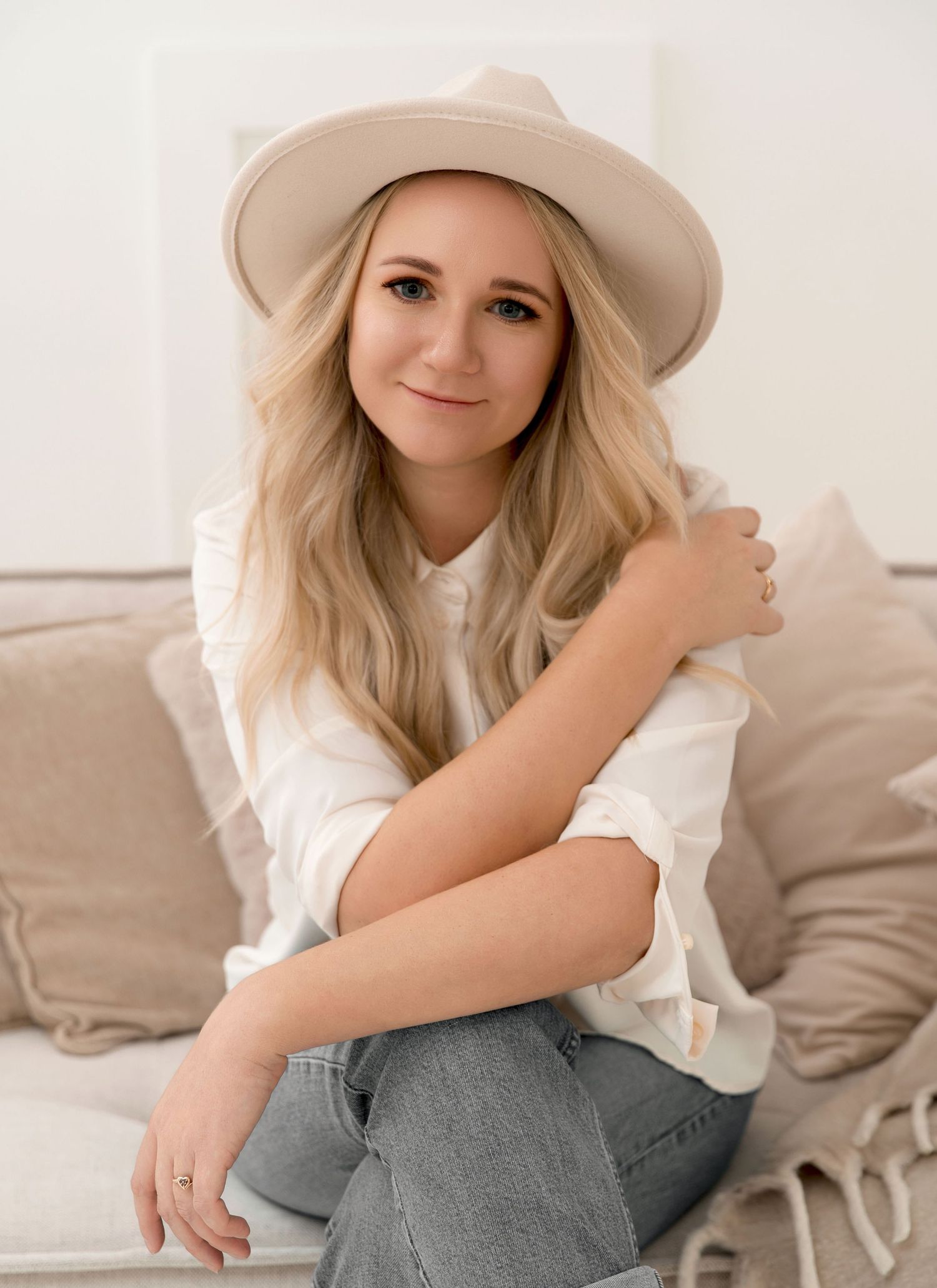 A person in a beige hat and white top poses casually on a couch with gray throw pillows in a bright room.
