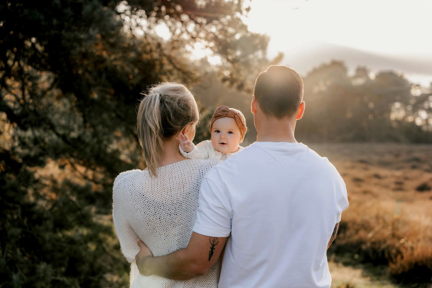 A family stands together in a golden field during sunset, backlit by warm sunlight filtering through distant trees.