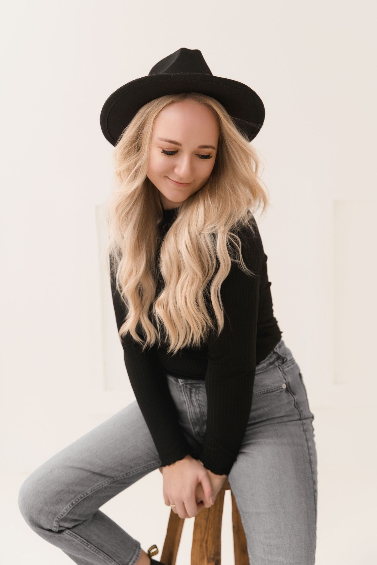 Stylish woman in black hat and sweater poses on wooden stool against light background in fashion photoshoot.