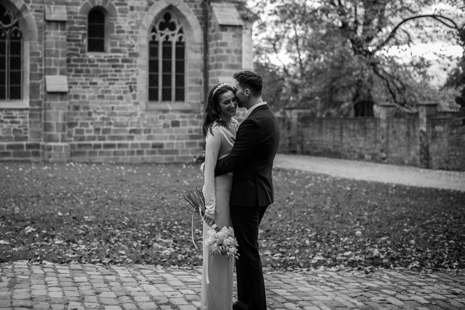 A black and white wedding portrait on church grounds with fallen leaves scattered around.