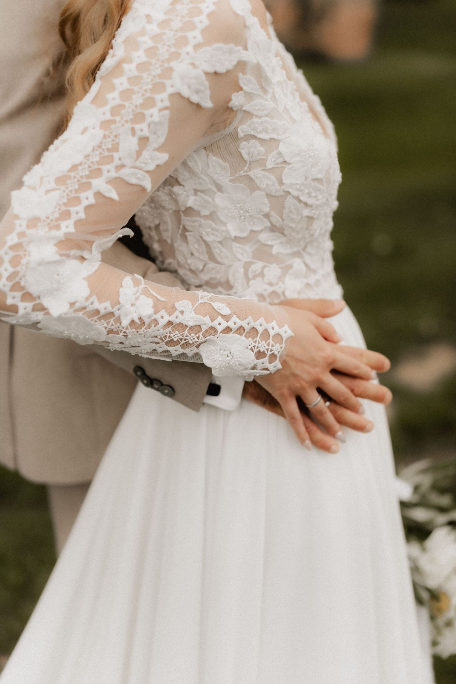 Close-up detail of delicate white lace sleeves and bodice of a wedding dress against a blurred outdoor background.