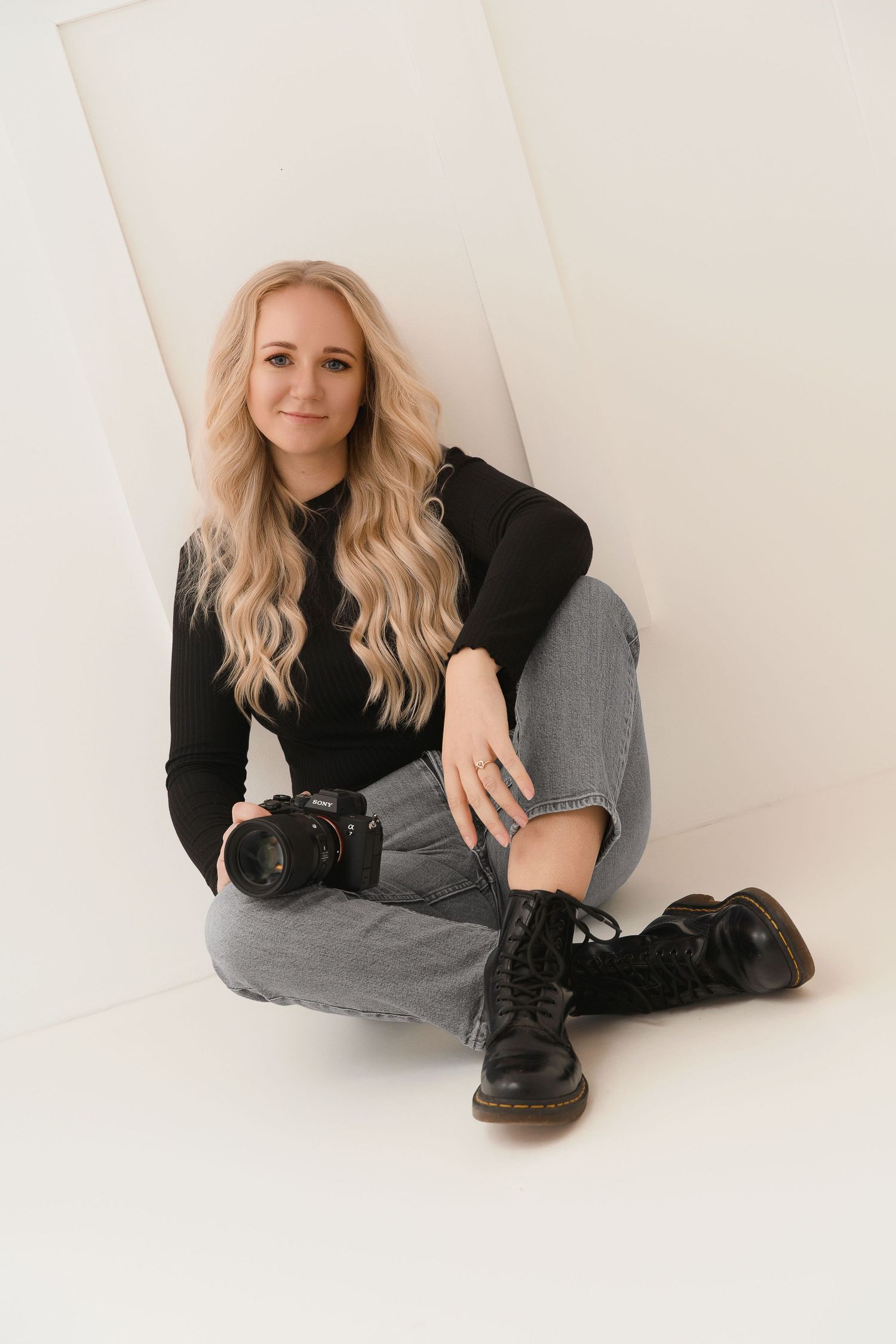 A photographer in black turtleneck and combat boots sits casually on white floor with camera resting nearby.