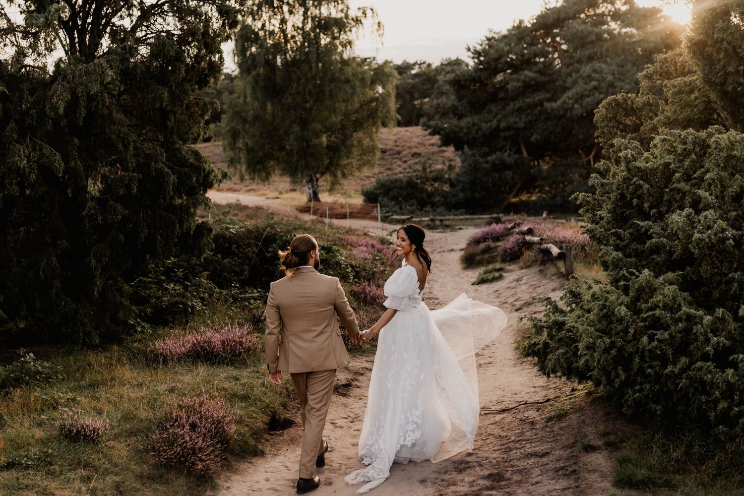 A wedding couple walking hand in hand along a scenic outdoor path at sunset.