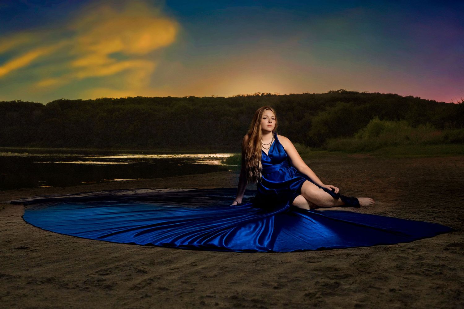 Woman in royal blue gown seated on sandy beach under colorful sunset sky.