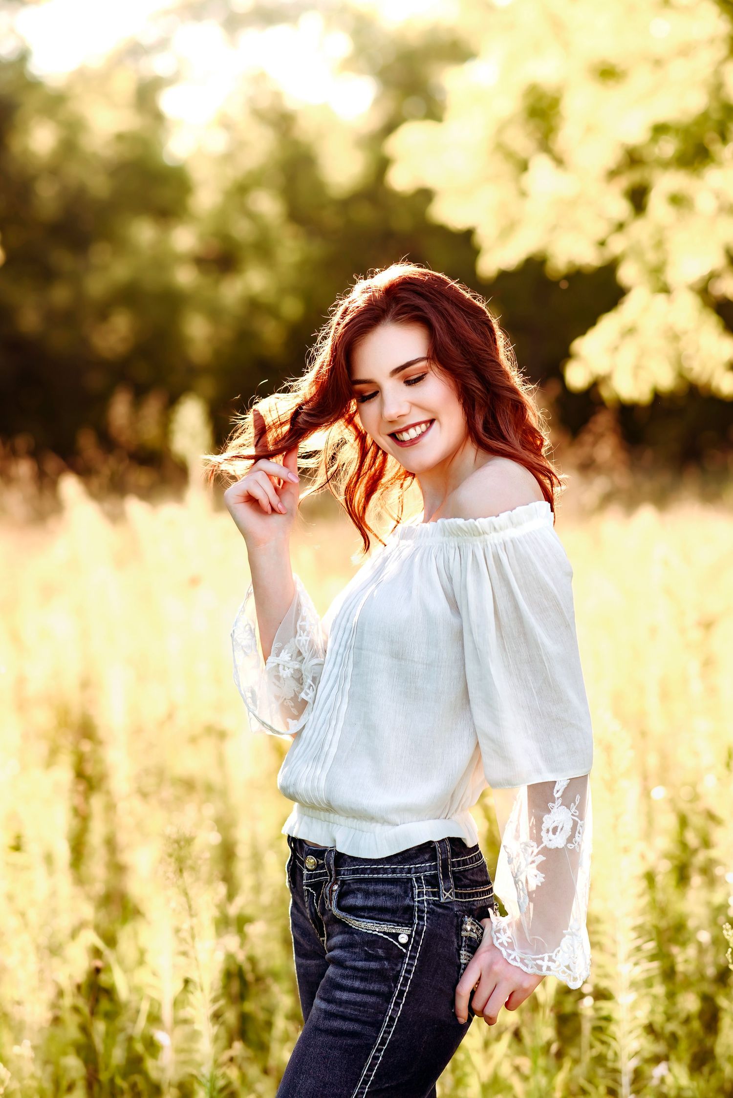 Smiling redhead woman in white off-shoulder blouse and jeans standing in a sunlit field.
