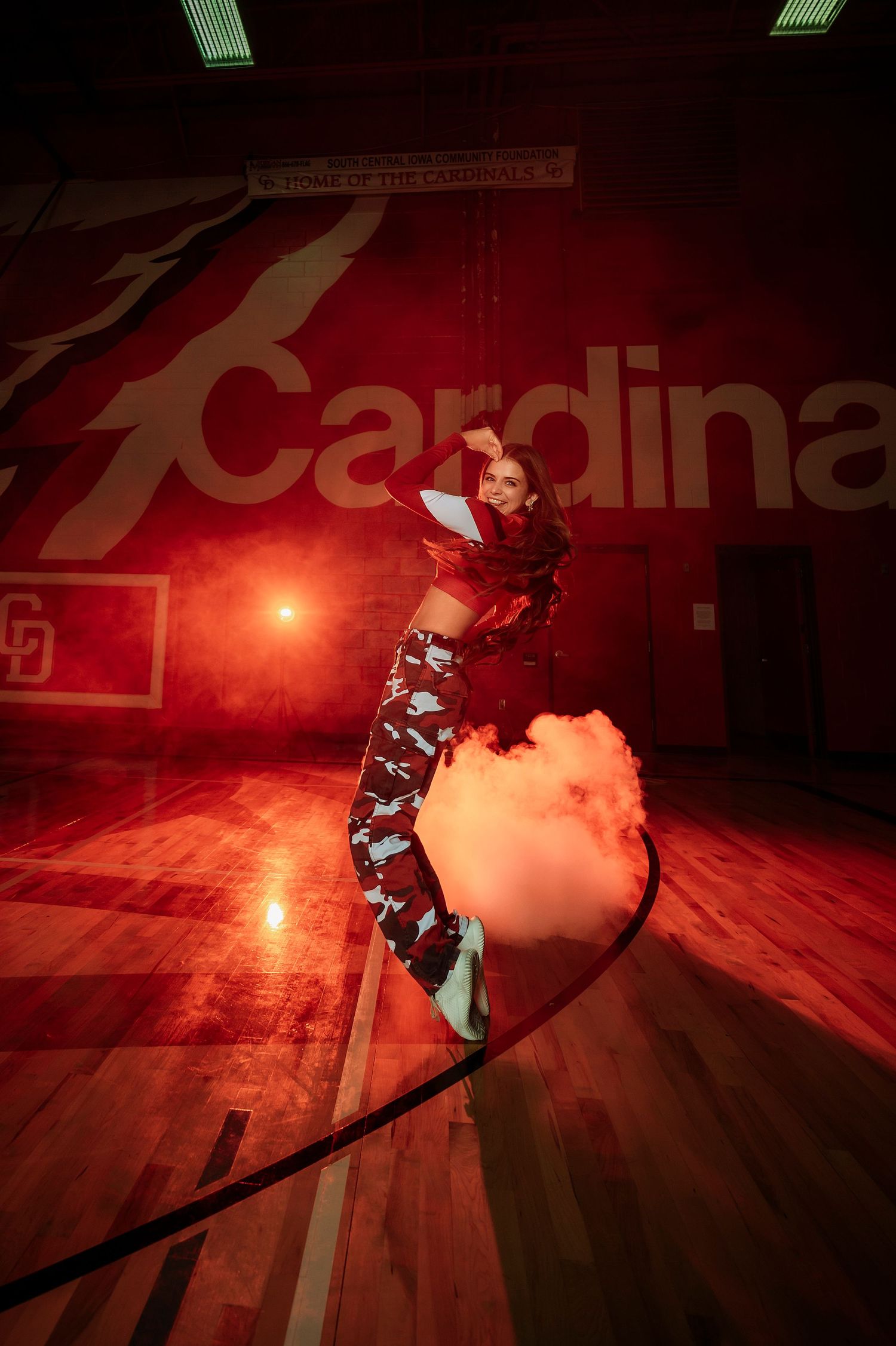 Female dancer jumps amid red smoke in Cardinals gymnasium with dramatic lighting.