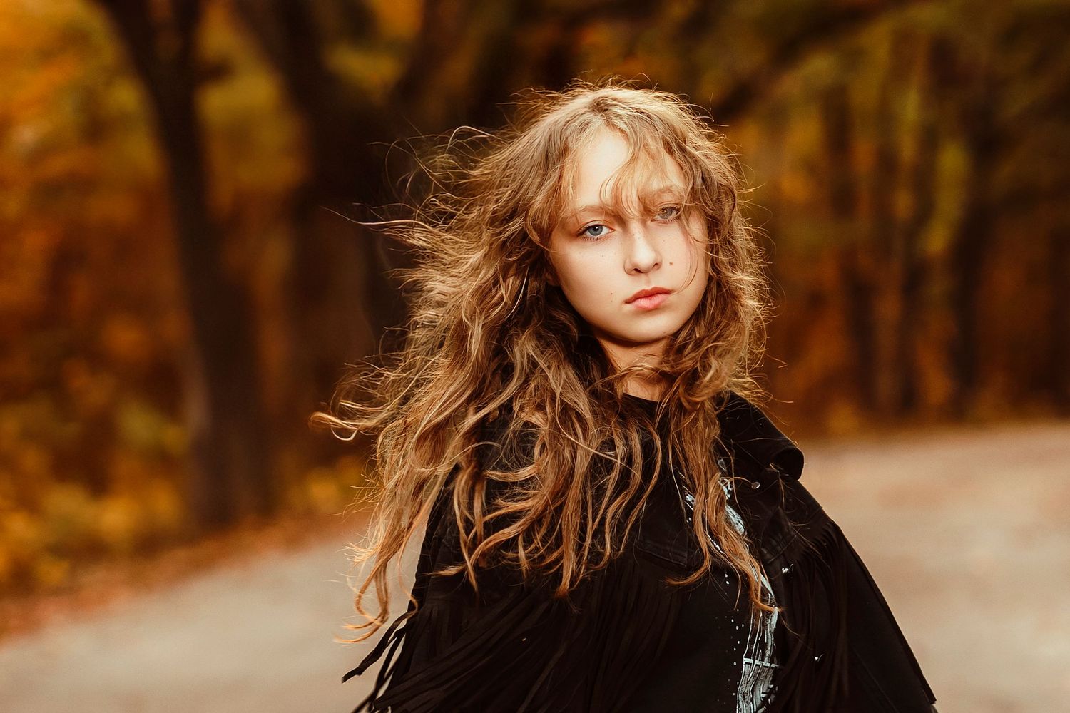 Young woman with windswept hair standing on an autumn road surrounded by colorful trees.