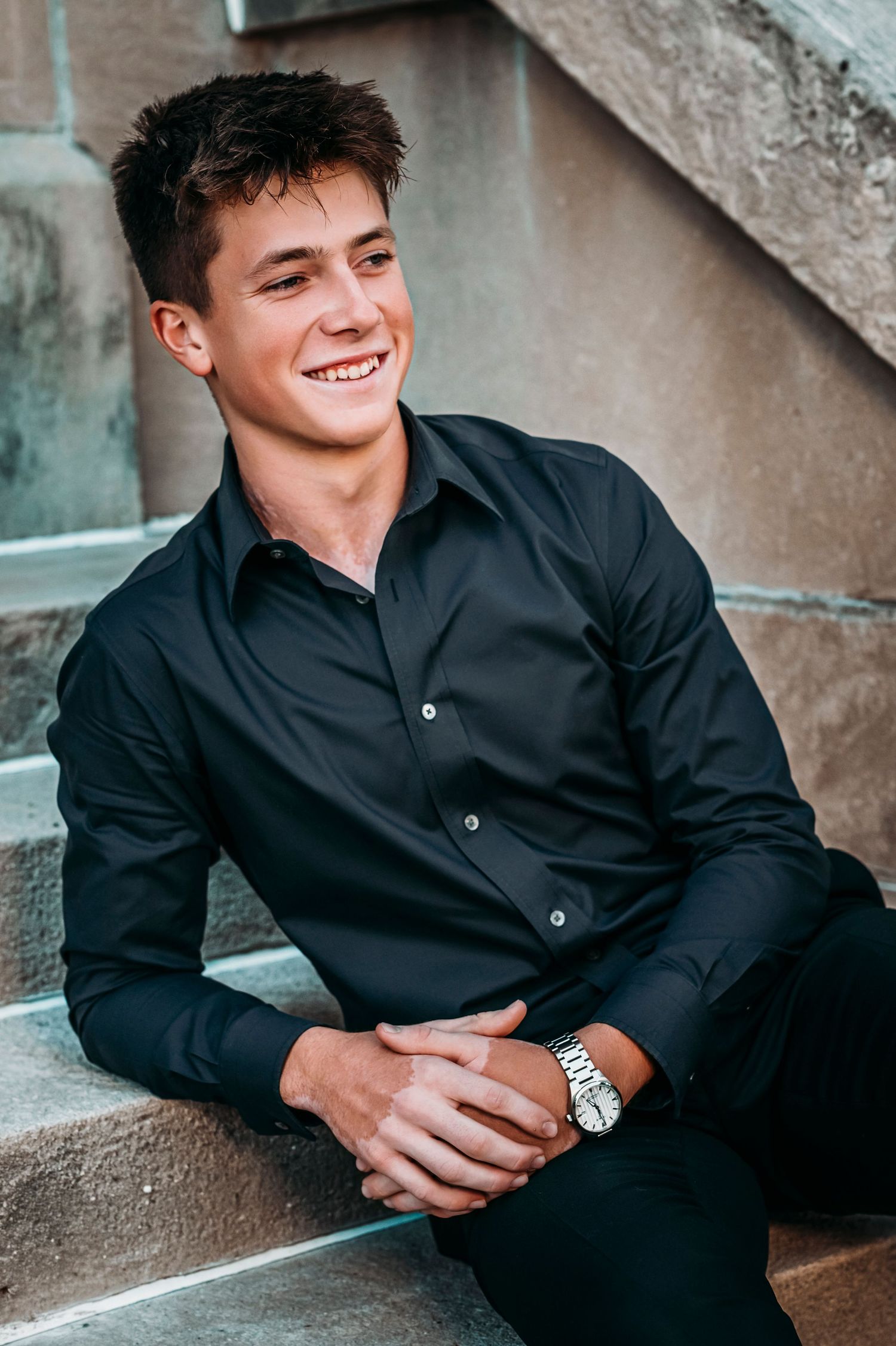 Smiling young man in black shirt sitting on stone steps wearing a silver watch.