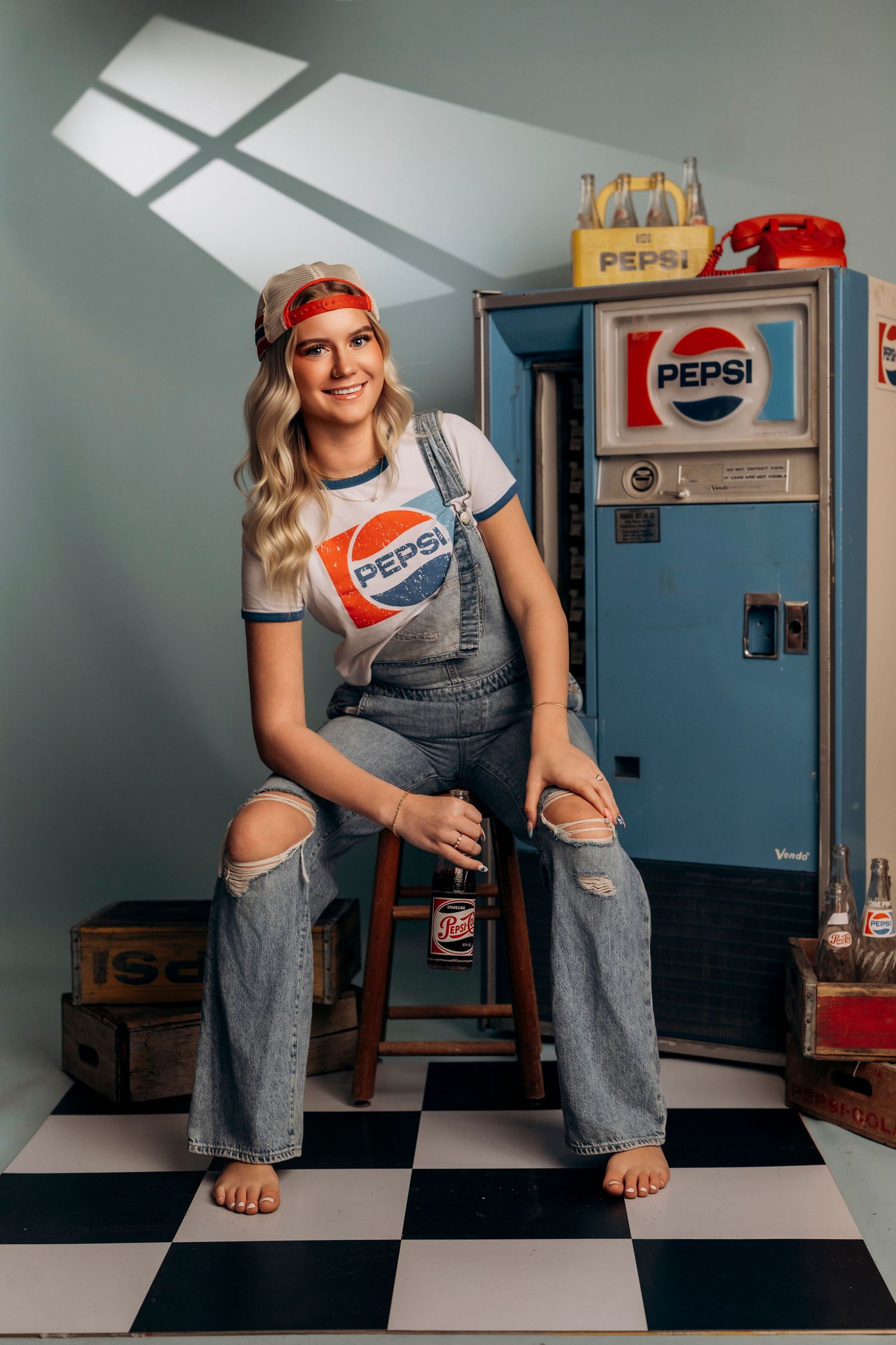 Smiling blonde woman in Pepsi t-shirt and ripped overalls sitting on stool near vintage Pepsi vending machine.