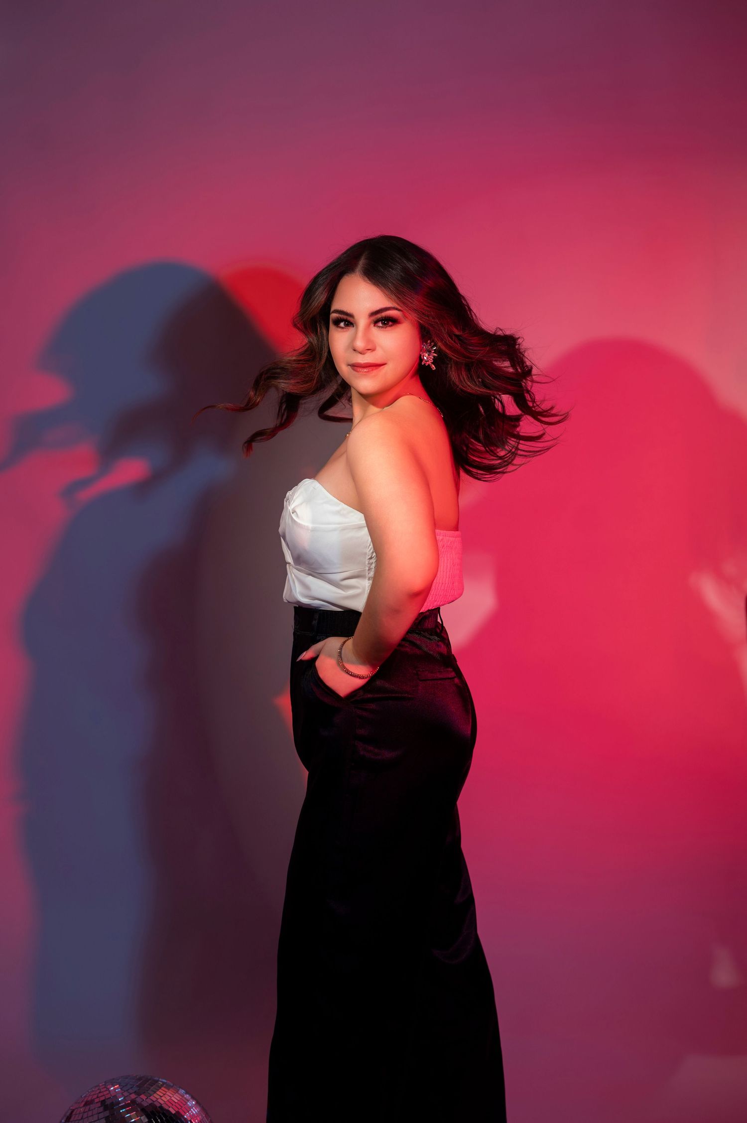Young woman in white top and black skirt posing against pink neon lit wall.