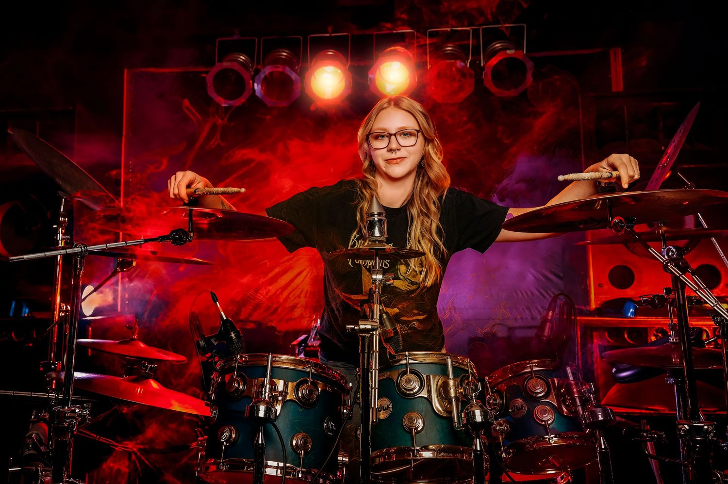 Young woman with glasses plays drums on stage with vibrant red concert lighting.