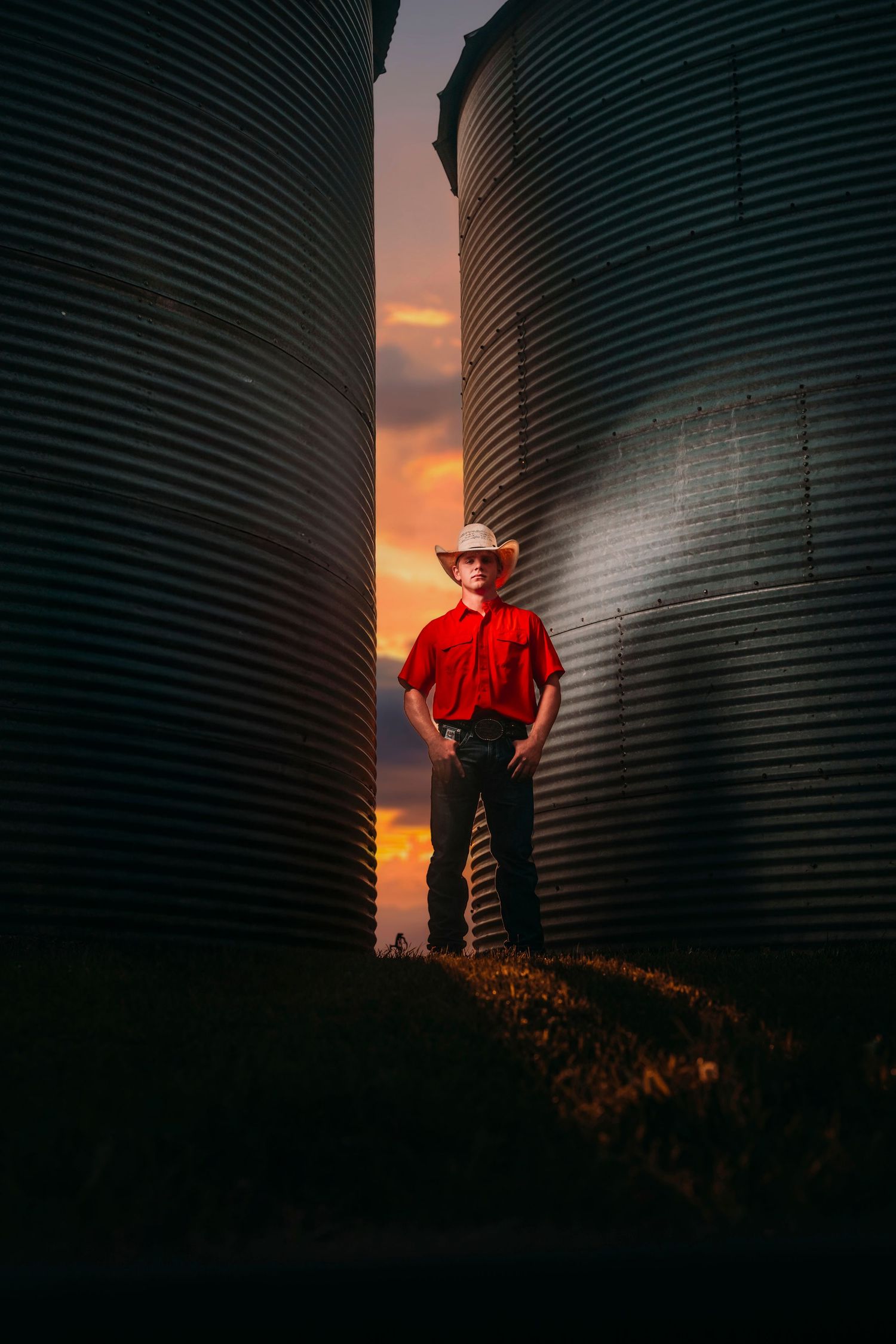 Young man in red shirt and cowboy hat standing between grain silos at sunset.