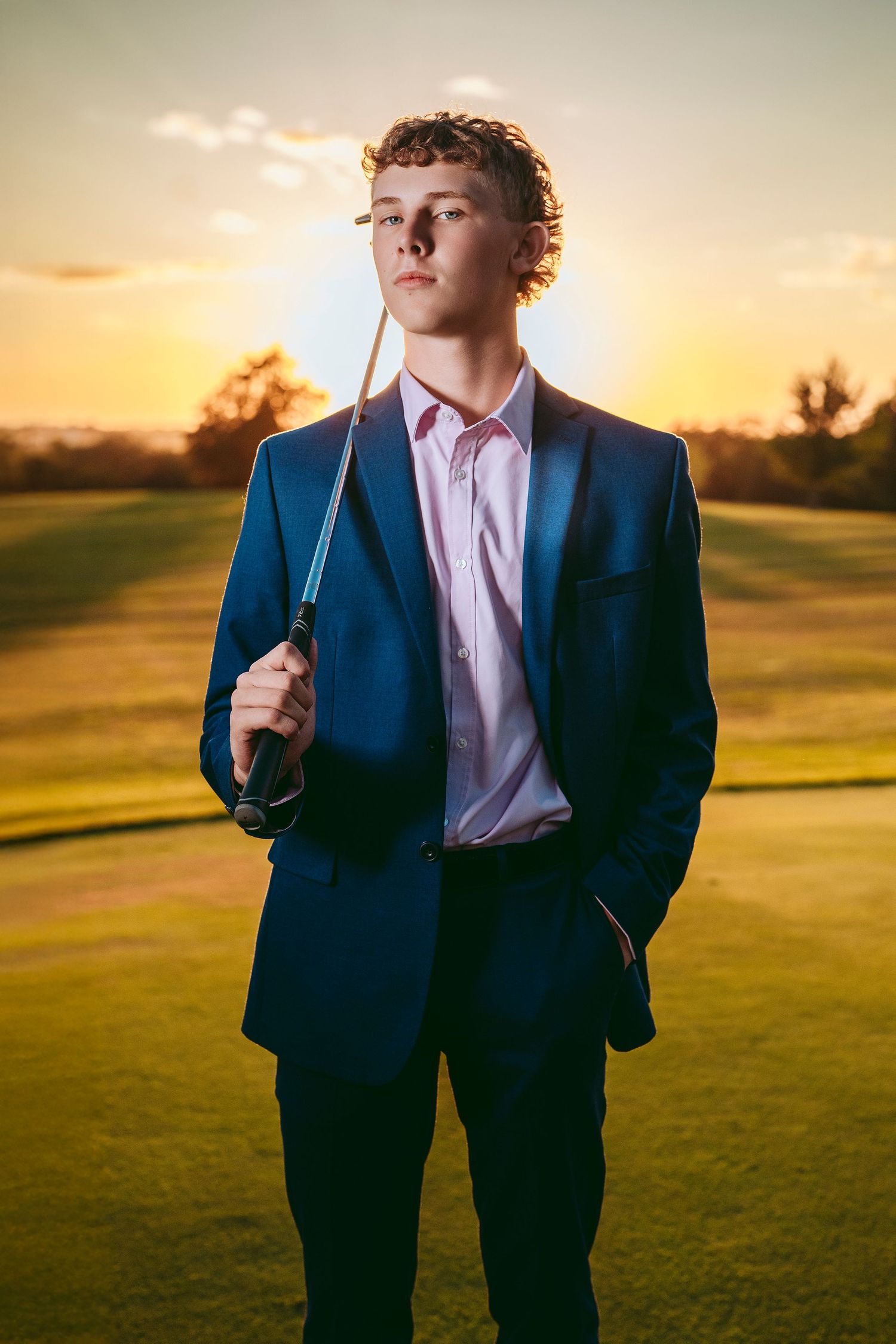 Young man in navy suit holding golf club on green course at golden sunset.