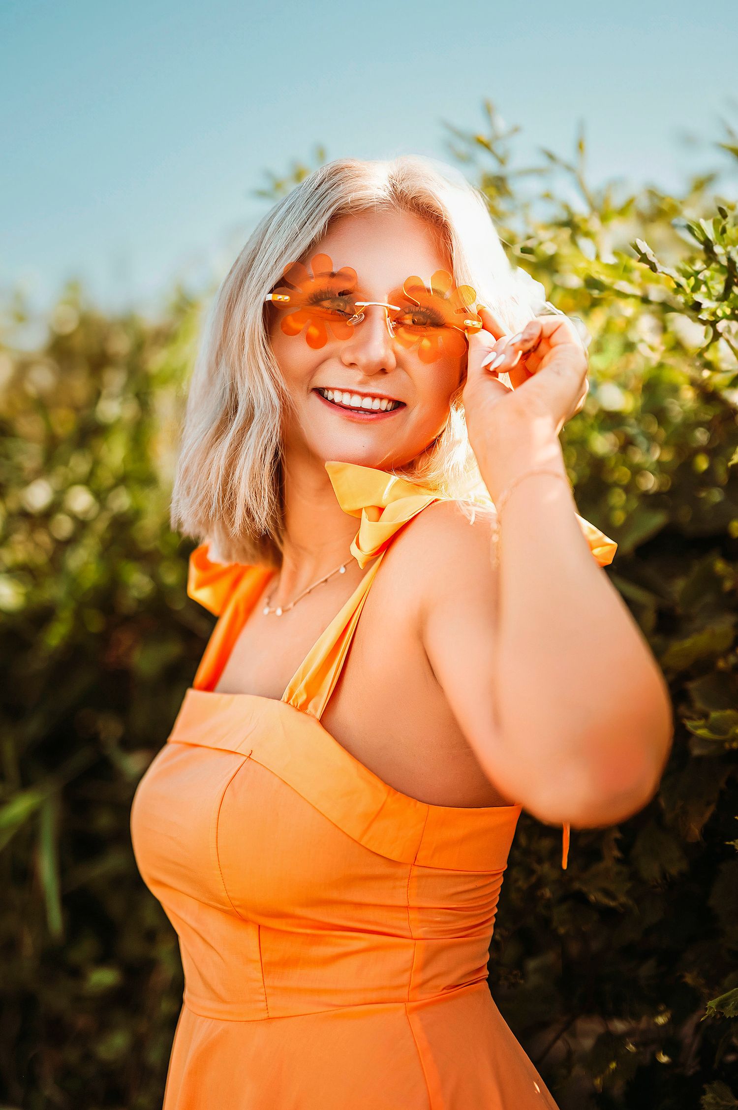 Smiling blonde woman in orange dress and sunglasses posing outdoors in bright sunlight.