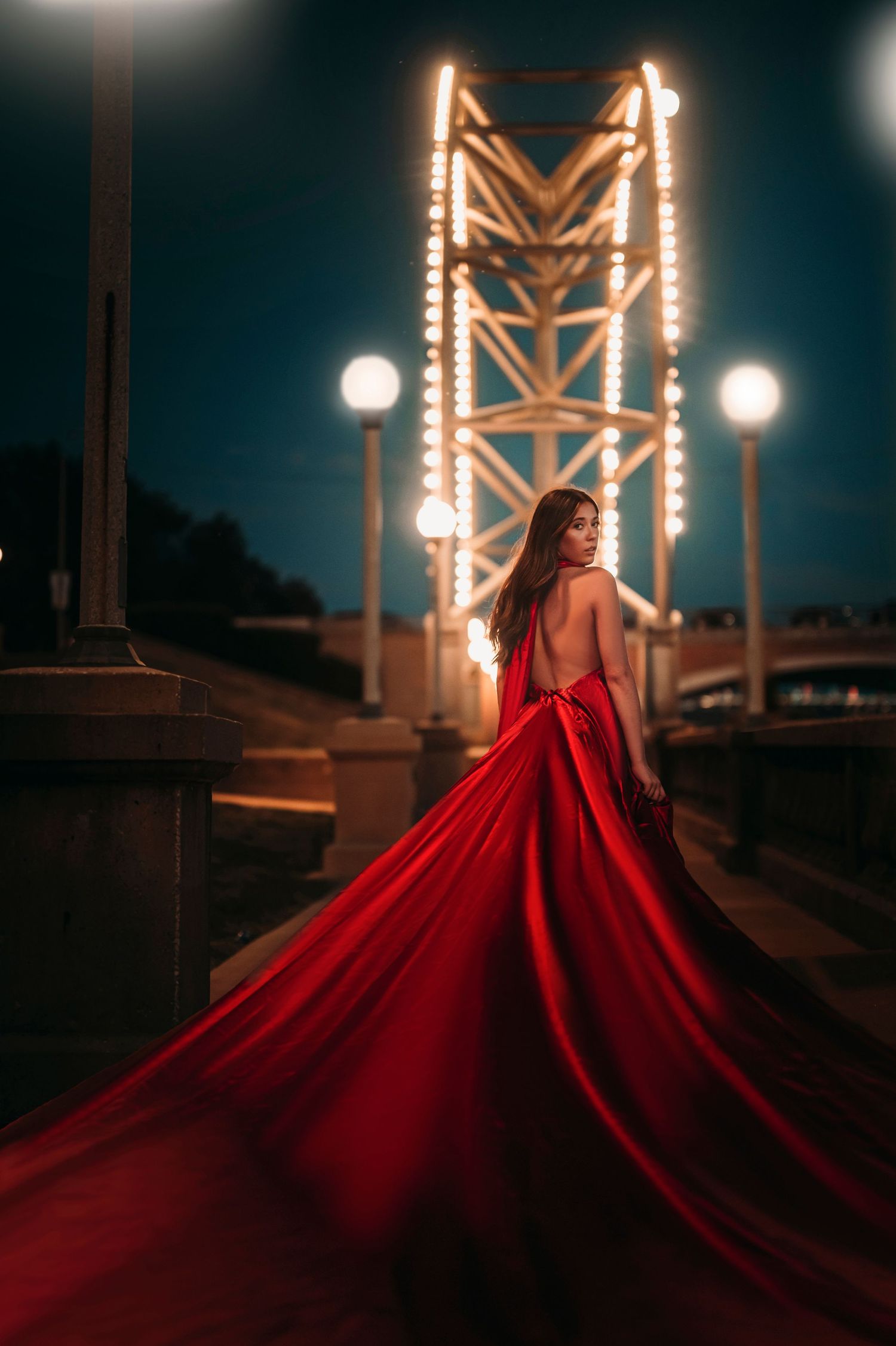Woman in flowing red gown with long train poses on bridge at night with glowing lights.