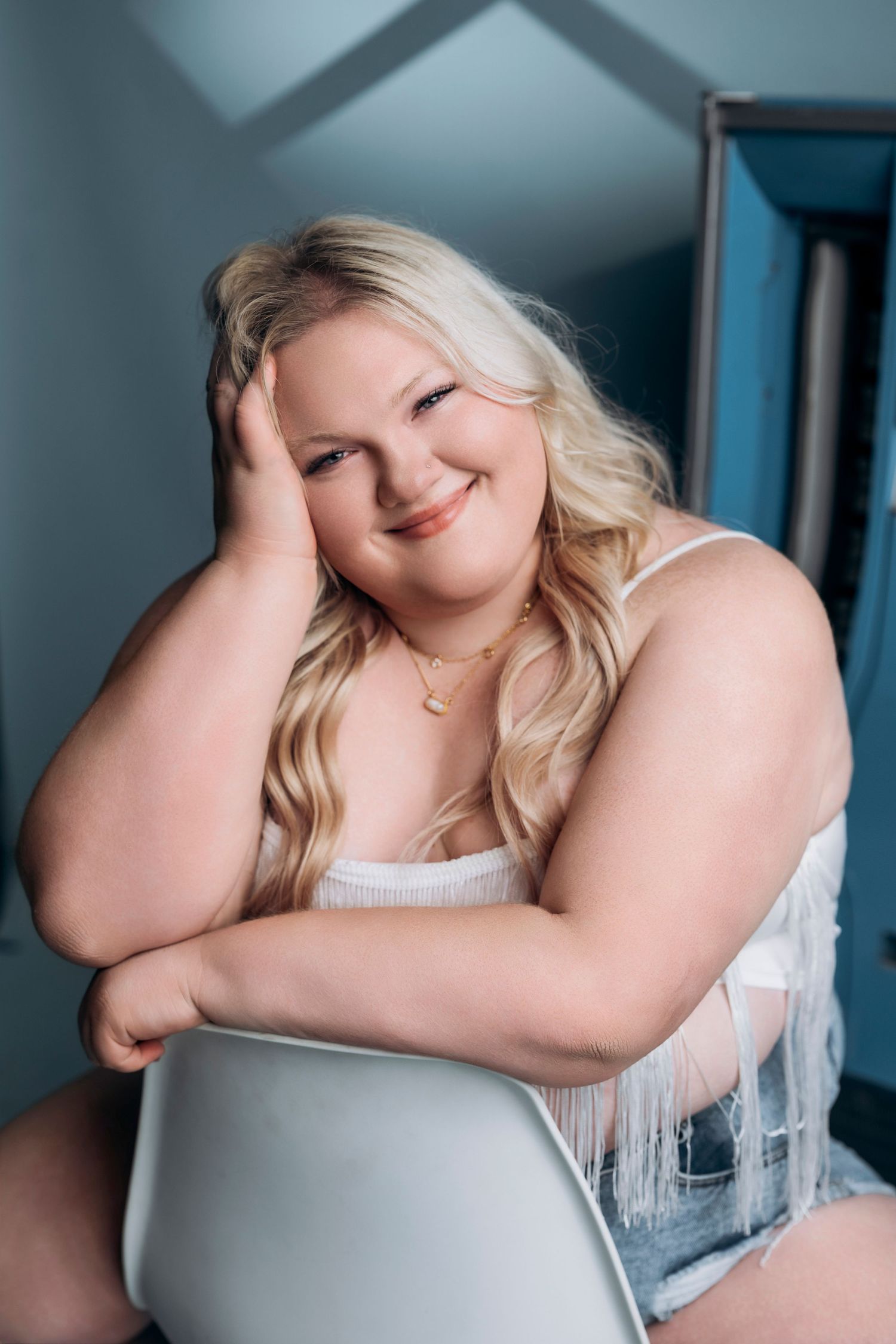 Smiling plus-size blonde woman in white fringe top and denim shorts sitting relaxed on a chair indoors. AMP Studio