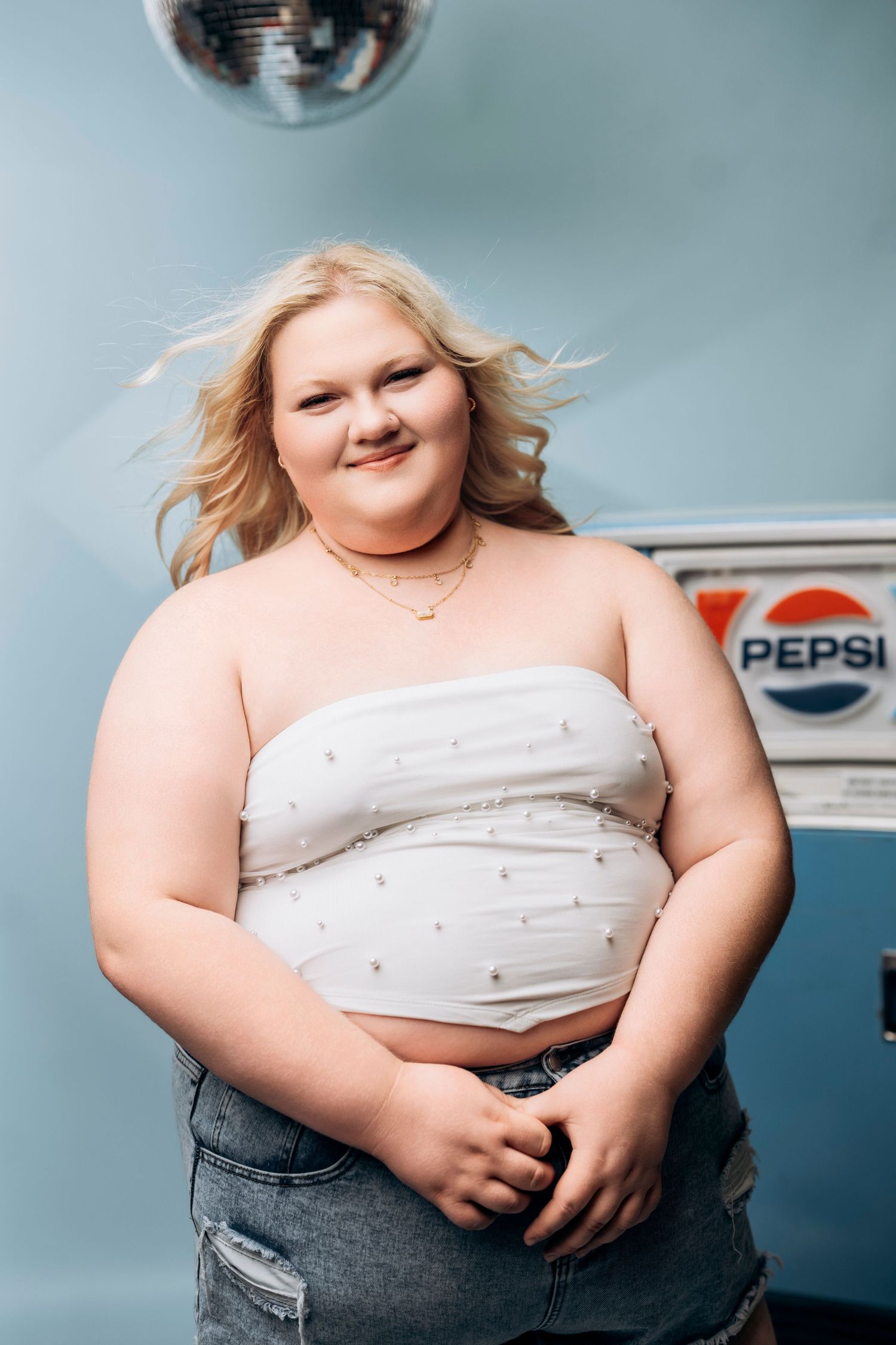 Smiling blonde woman in white tube top and ripped jeans posing near a vintage Pepsi machine. AMP Studio