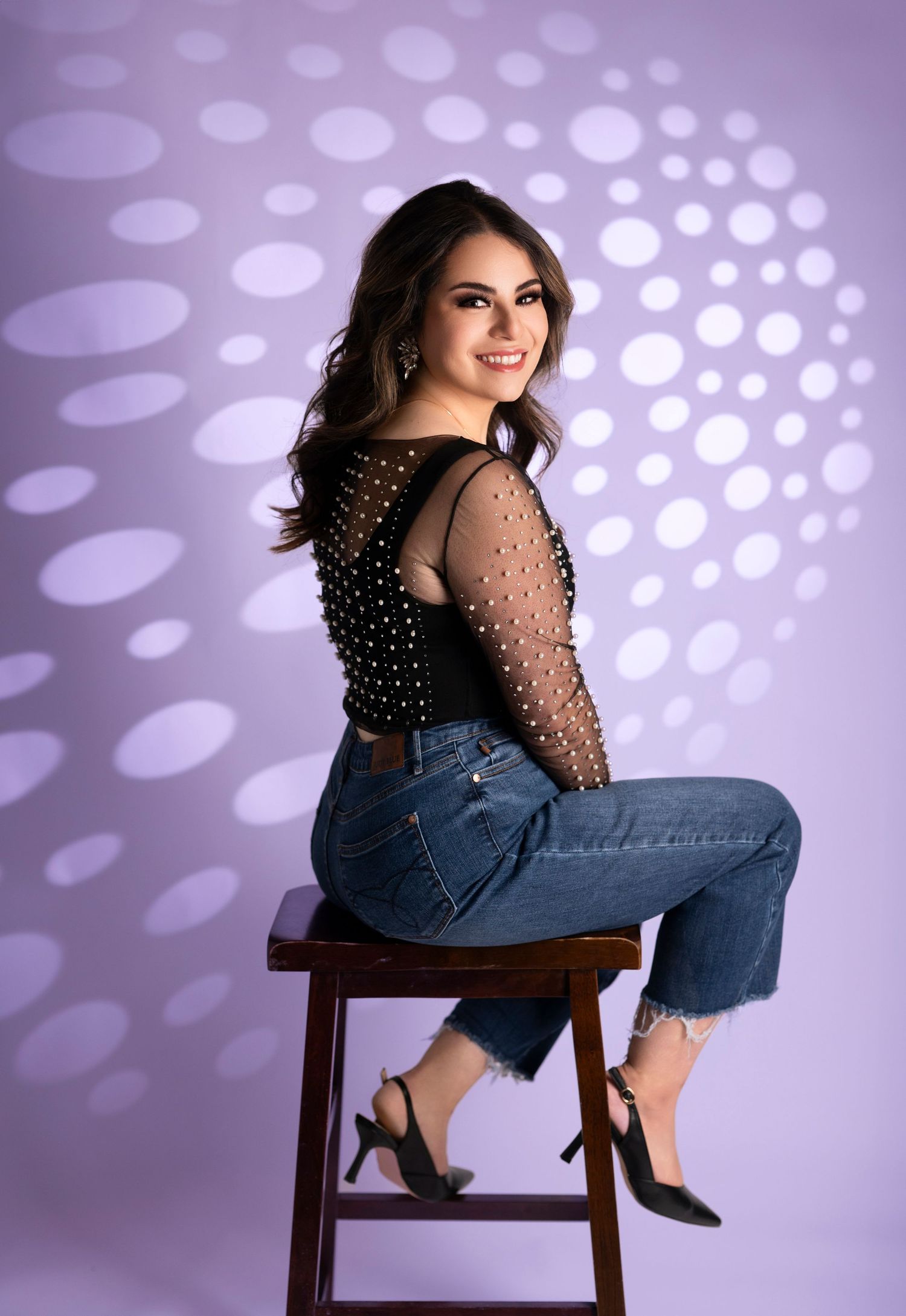 Smiling brunette woman in black pearl top seated on stool with purple dotted backdrop.